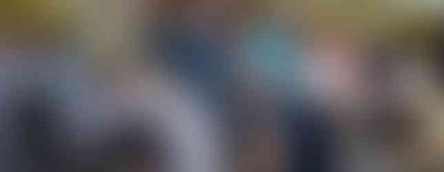 People seated outdoors in folding chairs, attentively listening. A woman with a walker sits beside others. Greenery in the background, clear sky.