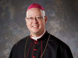Smiling man in clerical dress with a magenta zucchetto and gold chain against a textured gray background.
