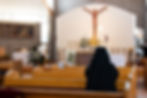 A nun in black habit prays in a church with wooden pews. A crucifix and altar with flowers and candles are in the background. Peaceful mood.