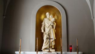 Statue of a robed figure holding a child in a gold alcove, surrounded by four candlesticks and a crucifix on a white altar. Calm mood.