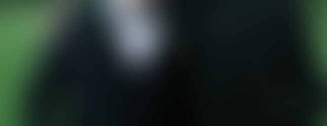 Two men in formal black suits, one in a clerical collar, smiling with arms around each other in a grassy outdoor setting.