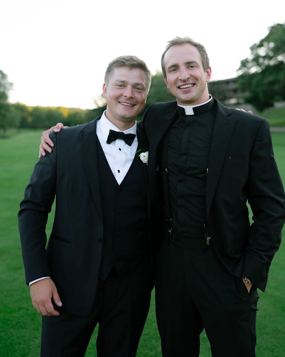 Two men in formal black suits, one in a clerical collar, smiling with arms around each other in a grassy outdoor setting.