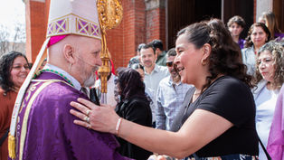 A person in a purple robe and mitre greets a smiling woman outside a brick building, surrounded by a group of people. Festive and friendly mood.