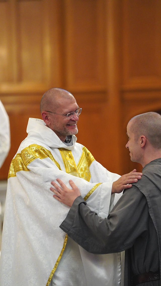 Priest in ornate white and gold robe smiles while embracing a man in gray, set in a warmly lit wooden interior, conveying joy.
