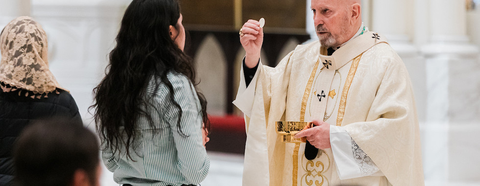 Priest in white robe giving communion to a woman in a church. Ornate altar in background. Calm, reverent atmosphere.