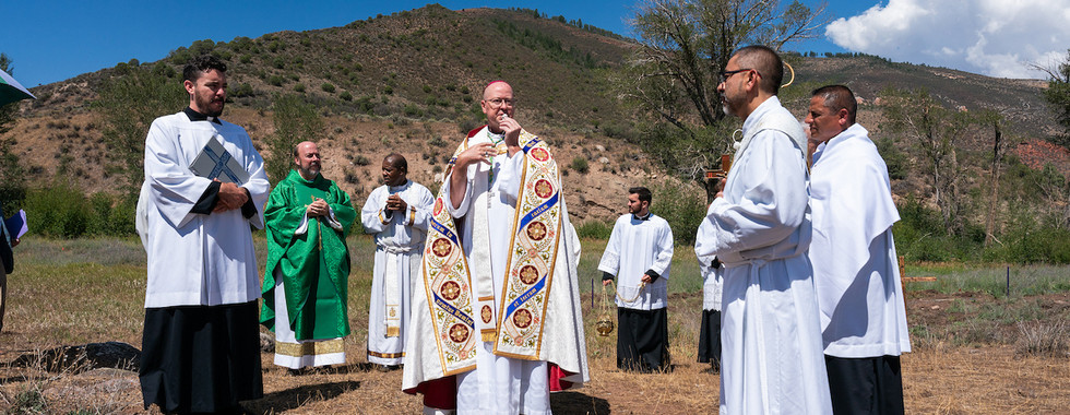 Priests in white and green vestments stand outdoors on a dry hillside under a blue sky, holding prayer items in a solemn ceremony