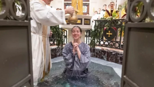 Priest pours water over a smiling woman in a baptismal font. Ornate church setting with gold accents and onlookers in the background.