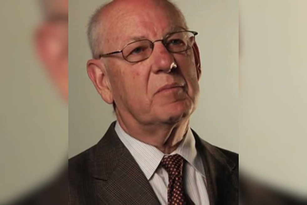 Portrait of an elderly man in a suit and glasses, looking serious against a plain beige background.