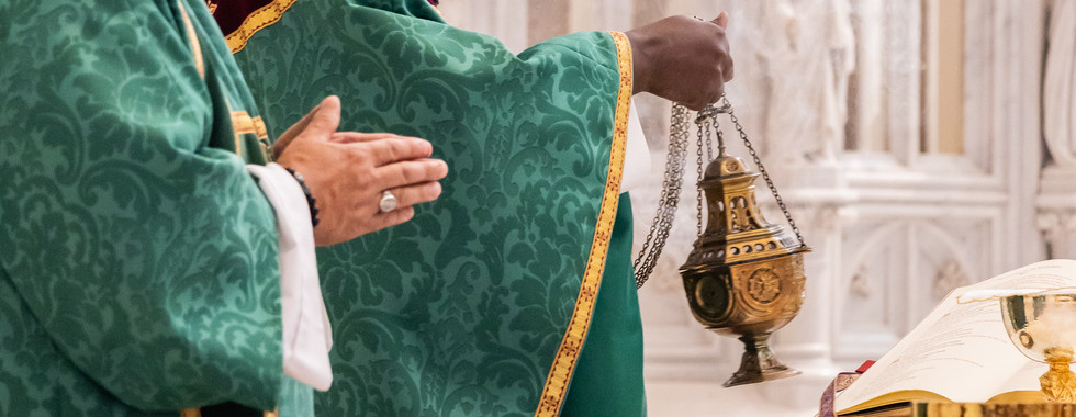 A priest and a deacon in green vestments at a church altar, one holding a hanging incense censer beside an open book, in a solemn ritual