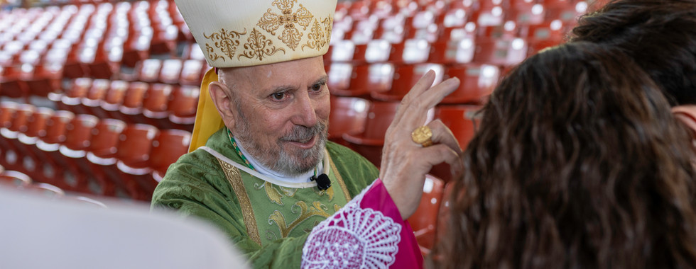 A bishop in green liturgical vestments and a mitre raises his right hand in a blessing gesture. He stands in front of rows of empty red seats in an indoor or covered venue.