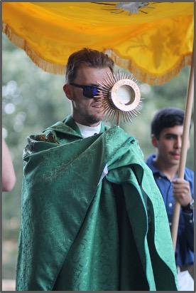 Man in green robe with sunglasses holds a religious symbol under a yellow canopy. Another person in blue shirt stands in background.