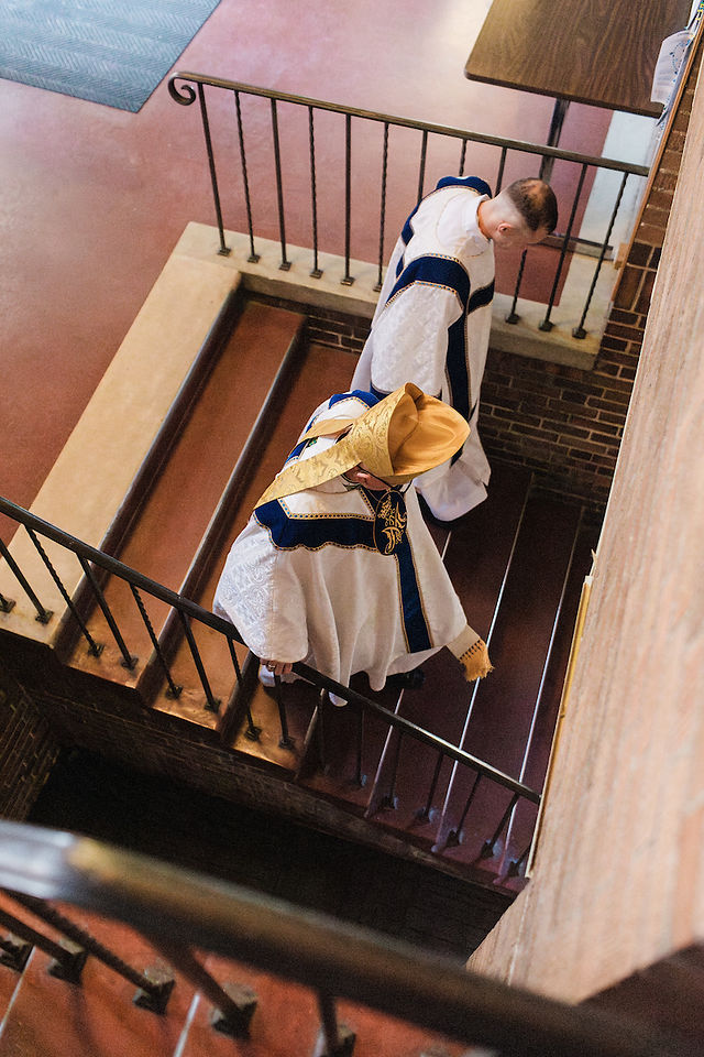 Two people in white and blue robes with yellow hats ascend a brick staircase in a dimly lit setting, suggesting a ceremonial mood.