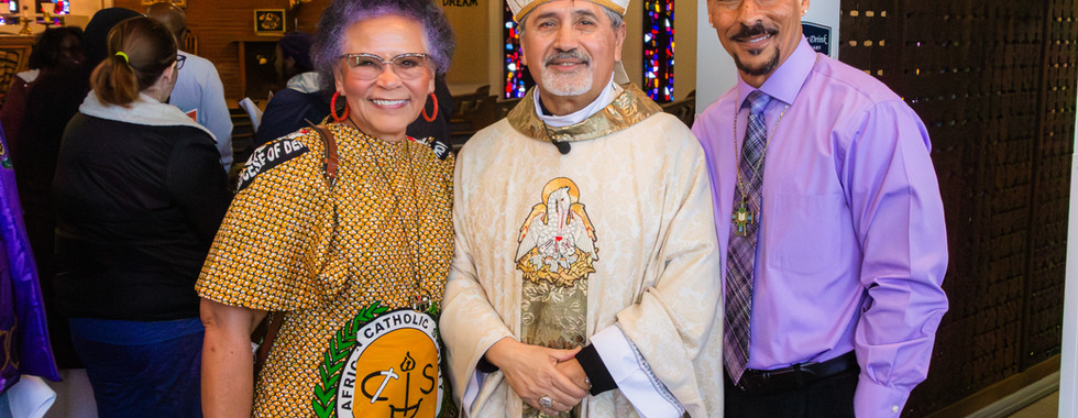 Three people pose together in a church with stained glass. One wears religious robes, another colorful attire. A banner reads "Live the Dream."