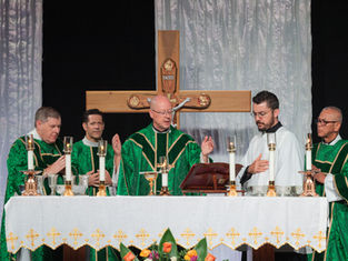 Sacerdotes celebran misa ante un gran crucifijo con INRI, altar con velas y mantel blanco; ambiente solemne.