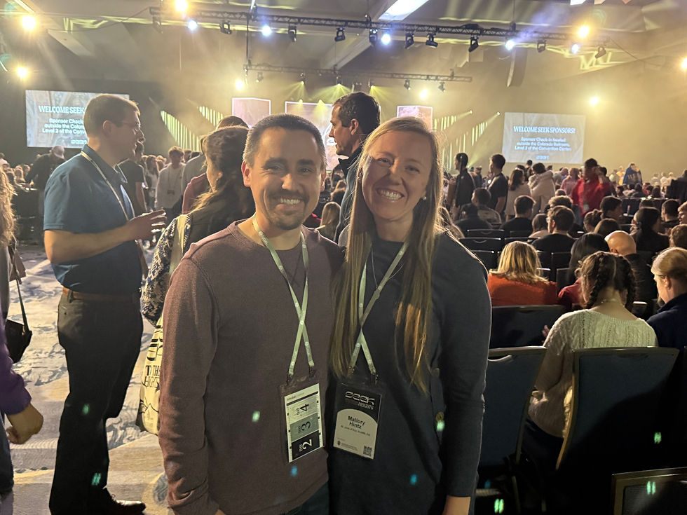 A smiling man and woman with name badges stand in a crowded conference hall. People sit and stand under bright lights, with welcoming signs.