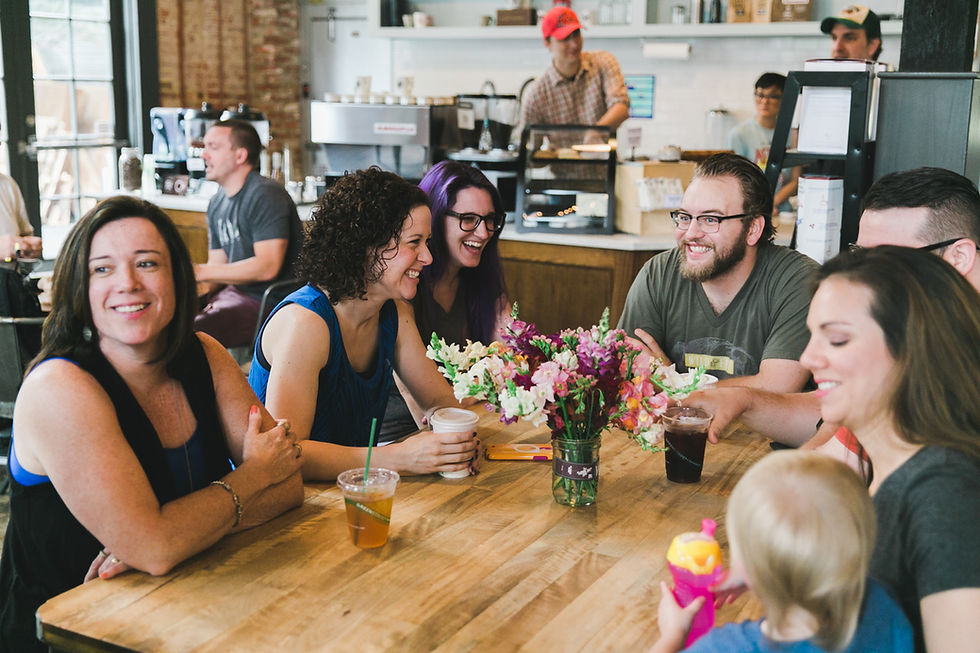 Group of people smiling and chatting around a table with drinks and a vase of flowers in a cozy café setting.