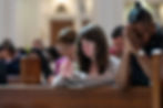 People praying in a church, eyes closed, hands clasped. Wooden pews and soft lighting create a serene atmosphere.