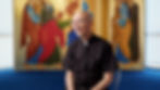 A person in clergy attire sits in front of a vibrant, detailed religious mural featuring figures in colorful robes. Bright, calm setting.