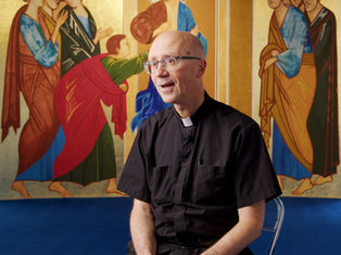 A person in clergy attire sits in front of a vibrant, detailed religious mural featuring figures in colorful robes. Bright, calm setting.