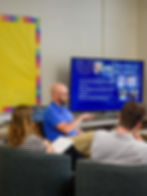 Three people in a discussion, one pointing at a screen titled "A Brief History of Good Shepherd." Bright yellow bulletin in the background.