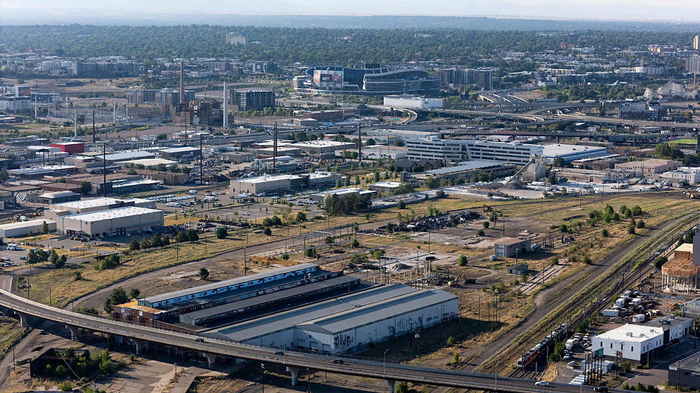 Aerial view of an industrial area with warehouses, rail lines, and scattered greenery. A distant stadium is visible under a clear sky.