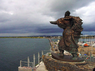 Bronze statue of a man pointing at sea, set on a stone platform overlooking a marina. Overcast sky and coastal road in the background.