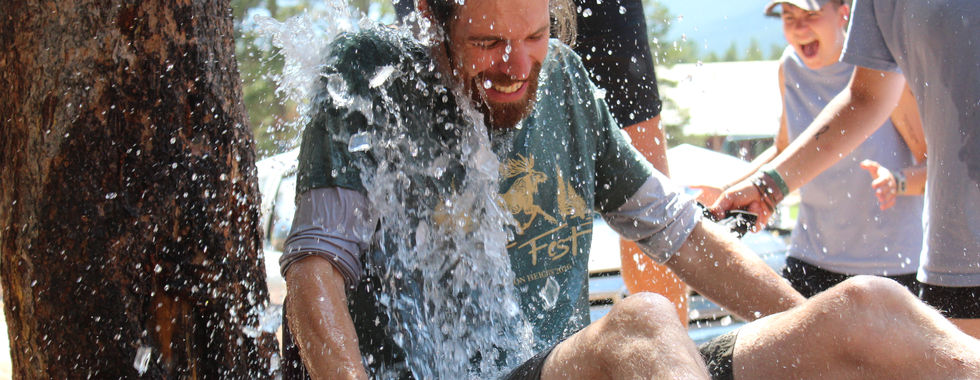 Laughing man gets doused with water in an outdoor tub while friends watch near a tree on a sunny day.