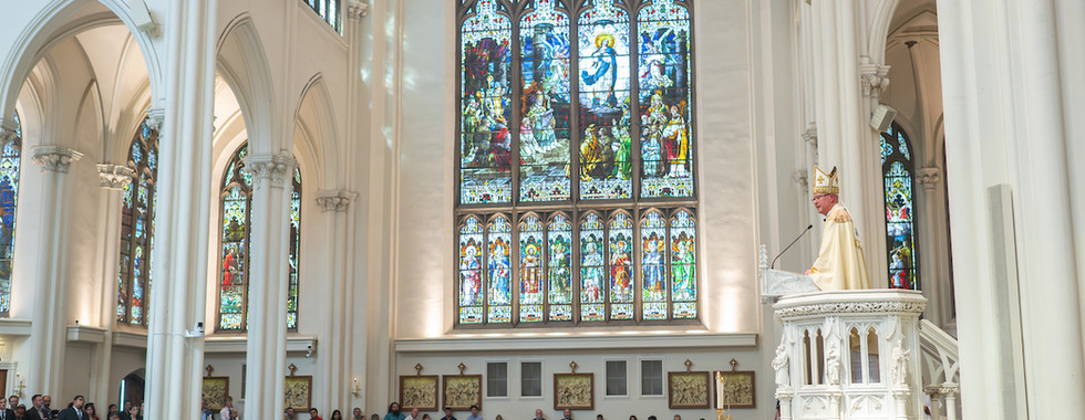 Cleric in white robes speaks at a pulpit in a cathedral. Stained glass windows and congregation fill the scene. Light and solemn mood.
