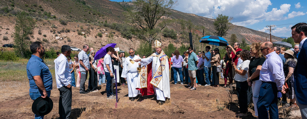 Bishop in ornate vestments blesses a crowd gathered in a dry mountain field, with umbrellas under a bright blue sky.