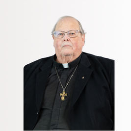Elderly clergyman in black attire with a cross necklace, neutral expression, light background.
