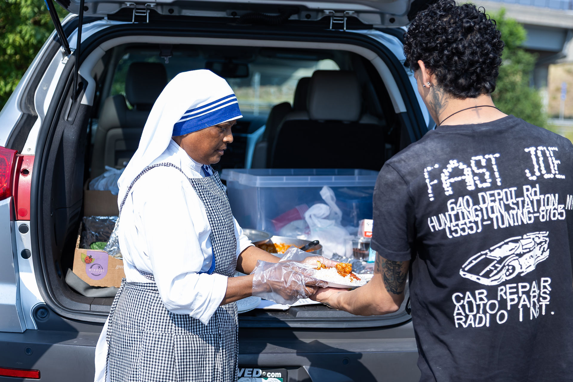 A woman in a headscarf serves food from a car trunk to a man in a "FAST JOE" shirt. Outdoor setting, bright day, giving mood.