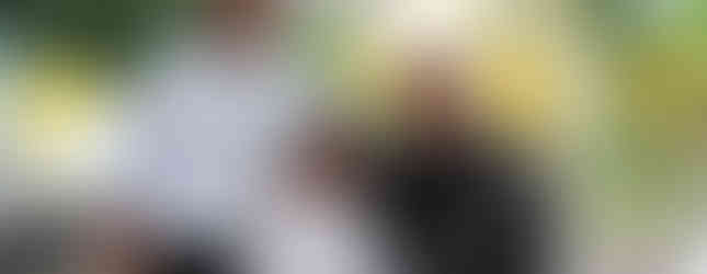 Three people smiling under a white canopy, with autumn trees in the background. One holds a long object. Casual outdoor setting.