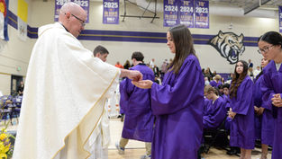 Priest gives communion to a student in purple graduation gown during a ceremony in a gym with banners and a tiger mural.