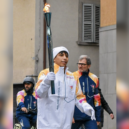 Person carrying a flaming torch, wearing "Milano Cortina 2026" jacket. Another person in similar attire nearby. Urban background, focused mood.