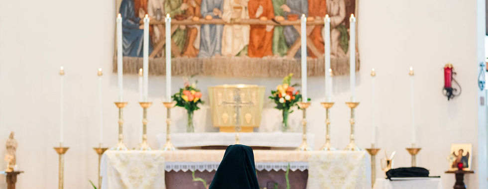 A religious sister kneels alone before the altar and crucifix in silent prayer during her vows ceremony.