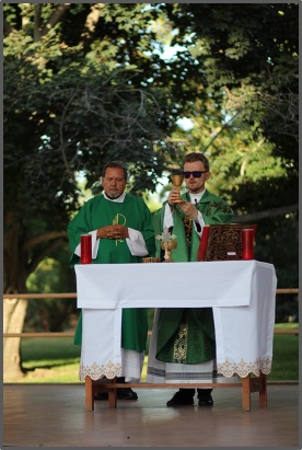 Two clergy in green vestments stand at an altar outdoors, holding a chalice and book. Trees are visible in the background, with a calm ambiance.