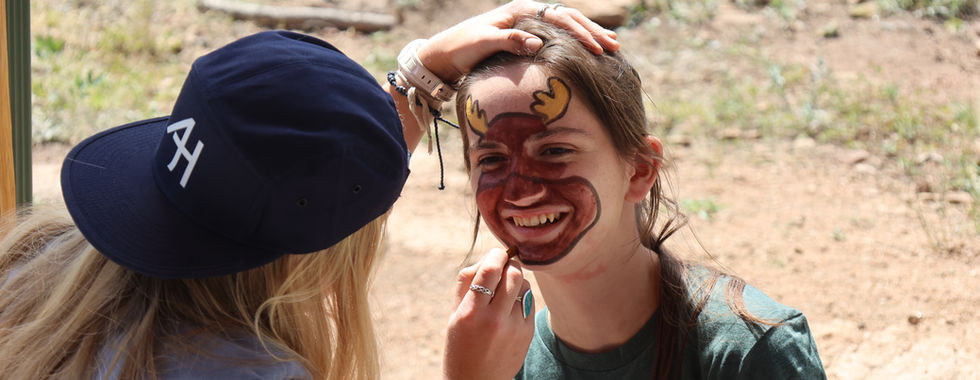 Girl paints a smiling teen’s face with deer antlers outdoors; navy cap with AH, rocky background, sunny scene.