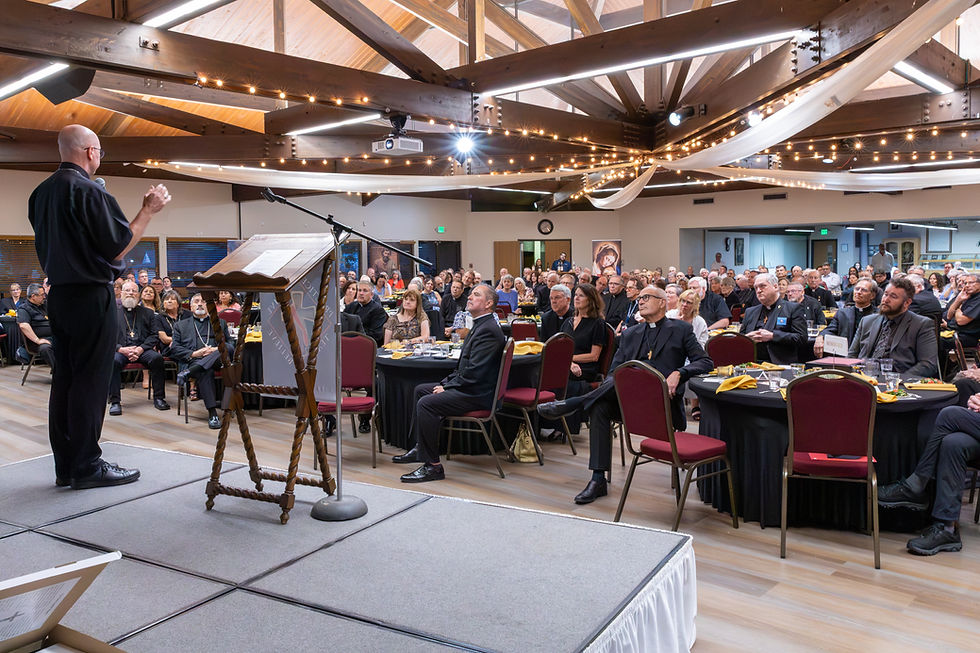 Priest speaking from a small stage to a large attentive audience at round tables in a warm, string-lit banquet hall.