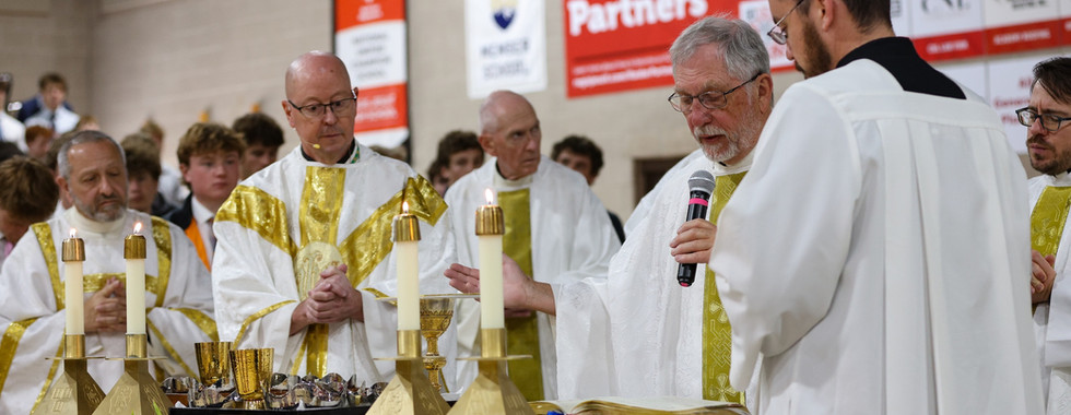 Priests and deacons gather around the altar during the Eucharistic Prayer at a Catholic Mass with candles, chalices and an open missal displayed.