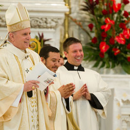 Clergymen in white, smiling and clapping, stand in an ornate church with red flowers in the background. One holds a paper. Joyful mood.