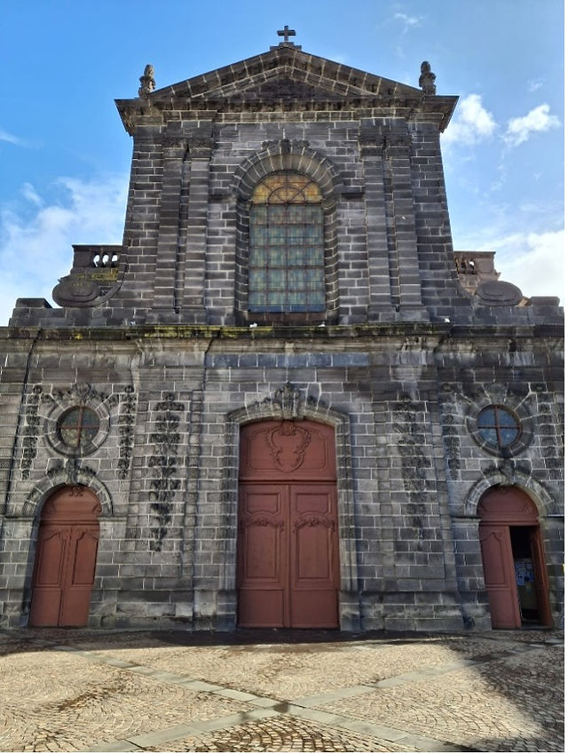 Stone church facade with large arched doors and stained glass window. Overhead, blue sky with few clouds. Cobblestone foreground.