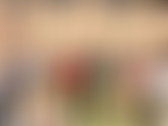 A group of eleven smiling men stand arm in arm on grass, with a brick building in the background. Bright, sunny day with casual attire.