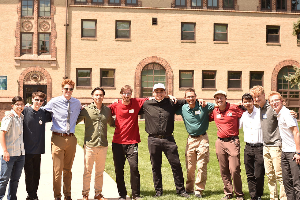 A group of eleven smiling men stand arm in arm on grass, with a brick building in the background. Bright, sunny day with casual attire.