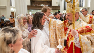 A bishop in ornate robes blesses a woman holding a candle in a church ceremony. People watch nearby, creating a solemn atmosphere.