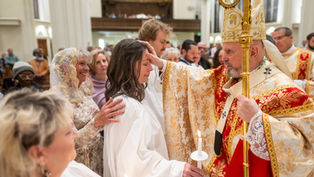 A bishop in ornate robes blesses a woman holding a candle in a church ceremony. People watch nearby, creating a solemn atmosphere.