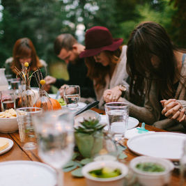 People sitting at a table outdoors, holding hands in prayer. Plates and glasses present, soft focus, greenery in the background.