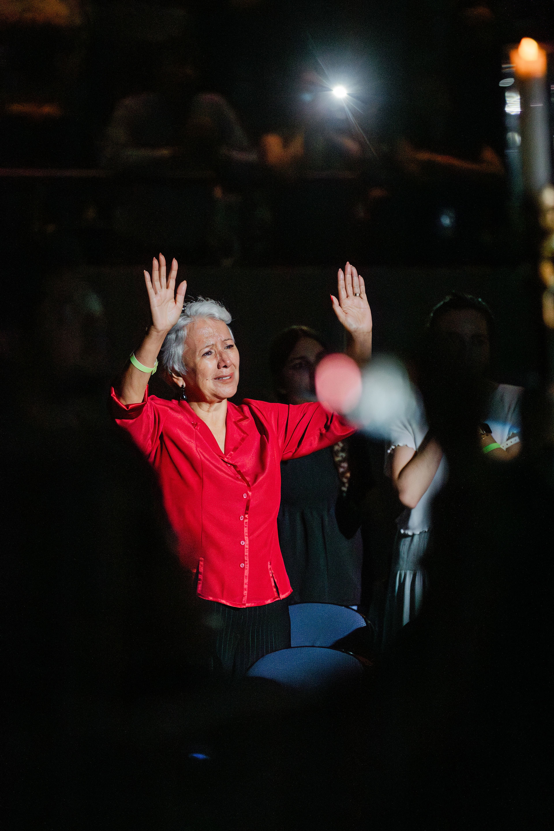 Elderly woman in a red shirt raises hands emotionally in a dark setting, surrounded by people. Flashlight illuminates her face.