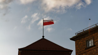 Polish flag on a building rooftop against a blue sky with scattered clouds; a bird flies in the distance. Calm, clear day.