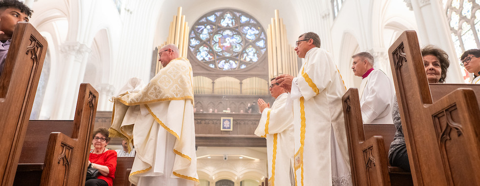 Clero de pie en oración en el pasillo de la catedral durante la liturgia, con la congregación sentada en los bancos.