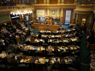 ista aérea de una asamblea legislativa con muchas personas de pie en una sala elegante. Sobresalen colores marrón y verde. Ambiente formal.